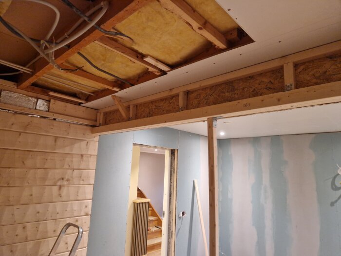 Renovering av ett rum med synliga träbjälkar i taket och väggar av panel och gips. En stege står mot en vägg.