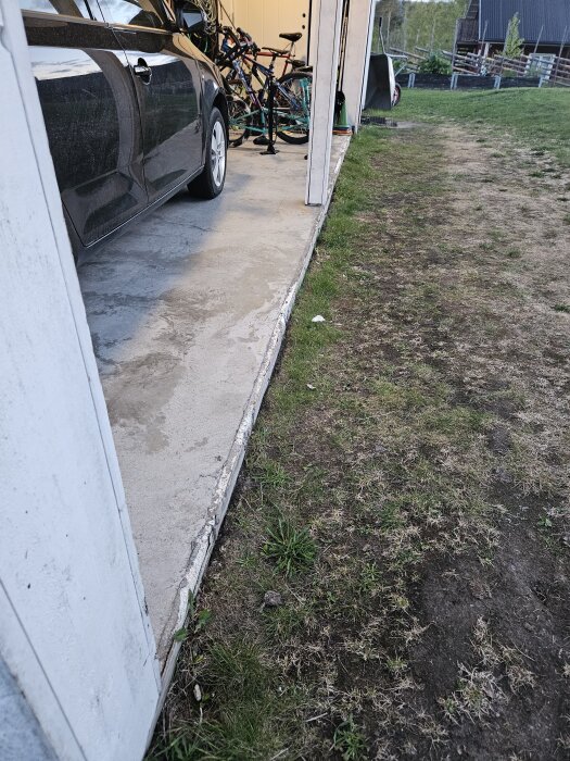 Carport med kant på bottenplattan delvis skadad. Bilar och cyklar syns i bakgrunden, gräsområde utanför plattan.