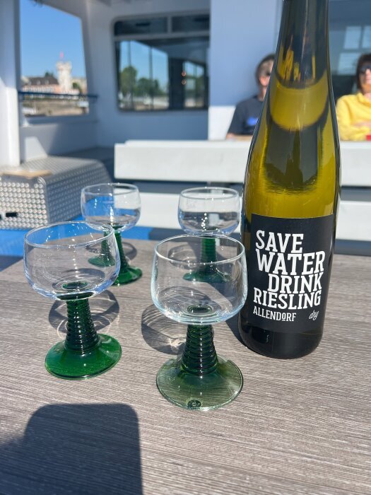 Vinflaska med texten "Save Water, Drink Riesling" står på ett bord tillsammans med fyra glas på en båt med personer i bakgrunden.