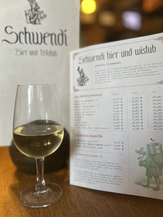 Glas med vitt vin bredvid en meny från Schwendi Bier und Wistub på ett träbord i en restaurangmiljö.