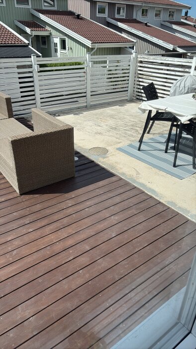 Uteplats med trätrall, betongplatta, möbler och vit staket med hus i bakgrunden under en solig dag.