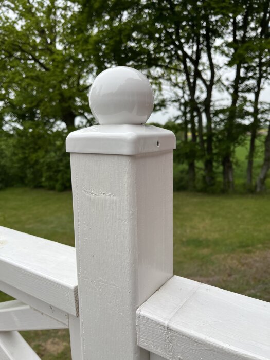 Vit stolphatt med rund topp monterad på balkongräcke i trä, omgiven av grönska i bakgrunden.