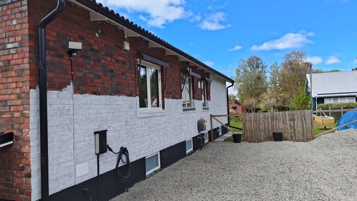 Delvis nymålad tegelvägg med en vit bas och två nyinstallerade fönster, omgiven av grus och trästaket.