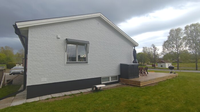 Nyrenoverad husvägg med vitt tegel, nytt fönster installerat. Träaltan med utemöbler och grill, omgivet av en grön gräsmatta.