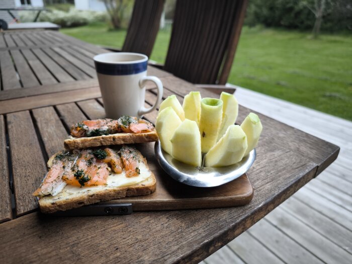 Gravlaxmackor, en kopp kaffe och skurna äpplen på ett träbord utomhus.