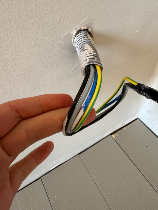 Hand håller i elektriska kablar med färgerna blå, gul-grön och svart som kommer ut ur ett rör i en vit vägg över ett grått golv.