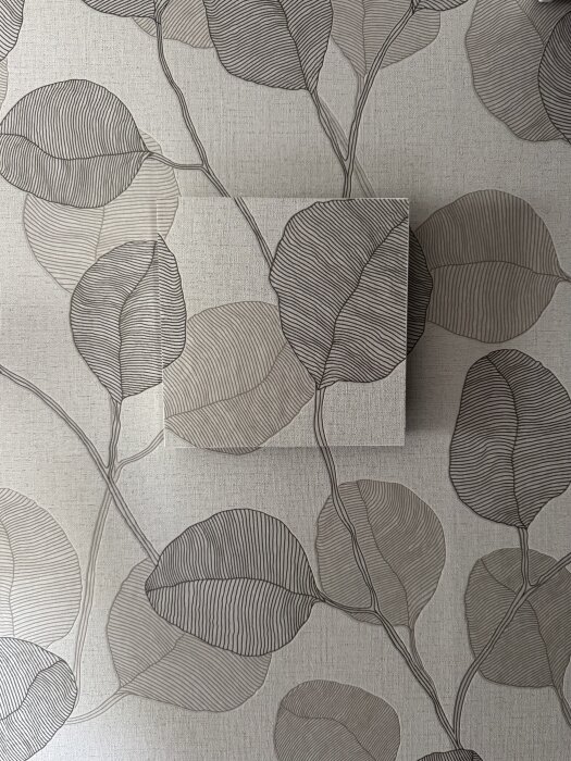 Mönstrad tapet med överlappande rektangulär panel, båda med samma lövdesign i grå nyanser.