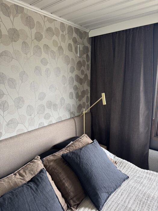 Sovrum med bladmönstrad tapet, mörka gardiner, säng med bruna och svarta kuddar, samt en guldfärgad läslampa.