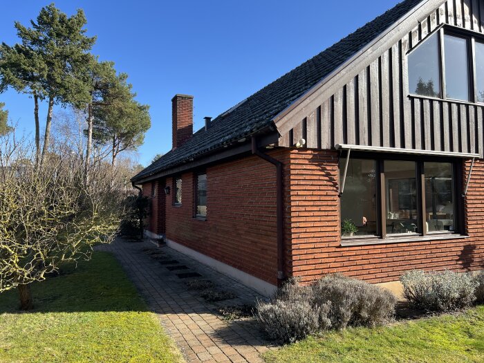 Rött tegelhus med nyrenoverad fasad och tak, stora fönster och trädgård i förgrunden; blå himmel och träd i bakgrunden.