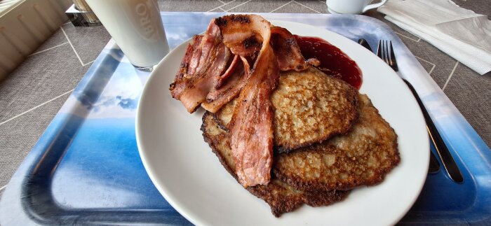 Raggmunk med fläsk och lingonsylt på en vit tallrik, serveras med ett glas mjölk, kniv och gaffel på en blå bricka.