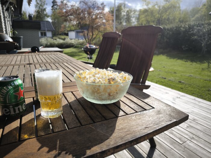 Popcorn och öl på ett träbord på en altan med en ölburk vid sidan, solig trädgård i bakgrunden.