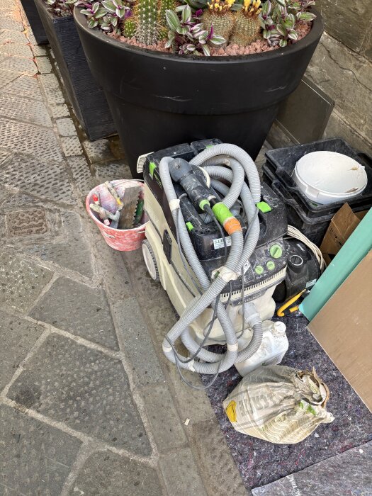 Verktyg och material för byggprojekt vid sidan av en stor blomkruka med kaktusar på en trottoar.