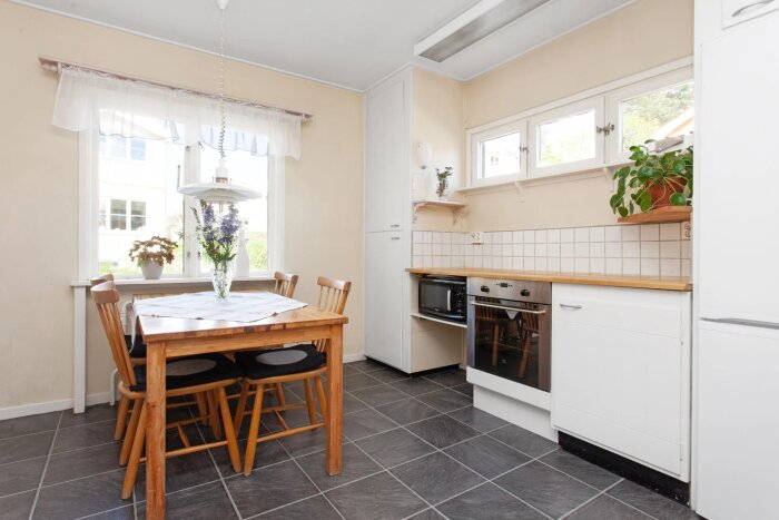 Ljust kök med träbord, fyra stolar, vita skåp och fönster. Blommor på bordet och hylla över diskbänken.