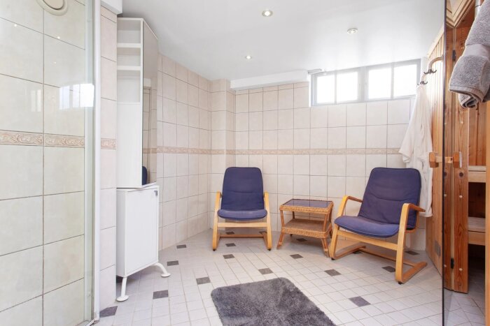 Två blå fåtöljer och ett bord i ett kaklat rum med dusch, matta, och handdukar på träkrokar.