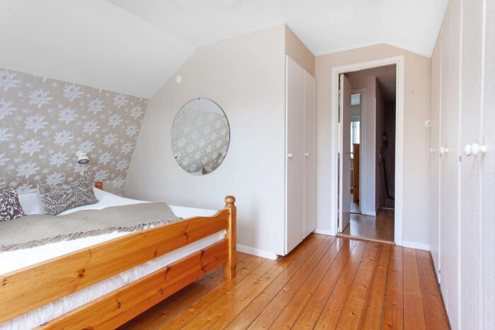 Sovrum med trägolv, blommig tapet och en träsäng; en rund spegel på väggen och öppna dörrar.