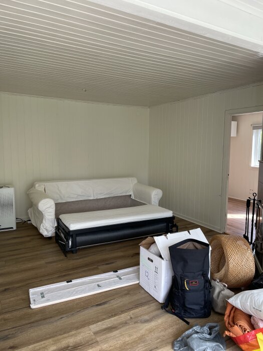 Bild på ett rum med vita väggpaneler och trätak. En vit soffa står vid väggen, omgiven av lådor och packning på ett trägolv.