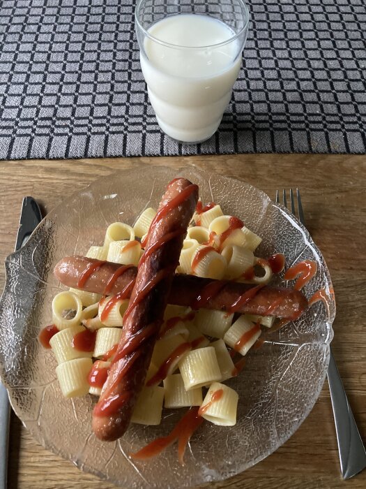 Tallrik med pasta och korv toppad med ketchup, serverad med ett glas mjölk på ett bord med bordstablett.