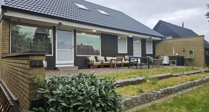 Exteriör av en tegelhusfasad med en terrass, sittmöbler, och grönska i förgrunden. Vattenslang rullar upp på väggen.