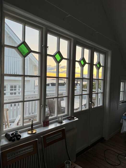 Fönster med spröjs och gröna rutor i sekelskifteshus, med utsikt över terrass och solnedgång vid havet.