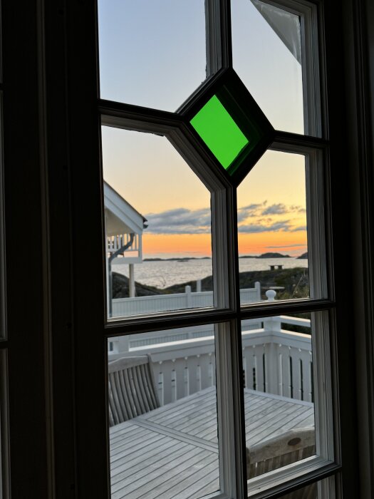 Ett spröjsat fönster i ett hus vid västkusten med utsikt över en träaltan och havet i solnedgången.