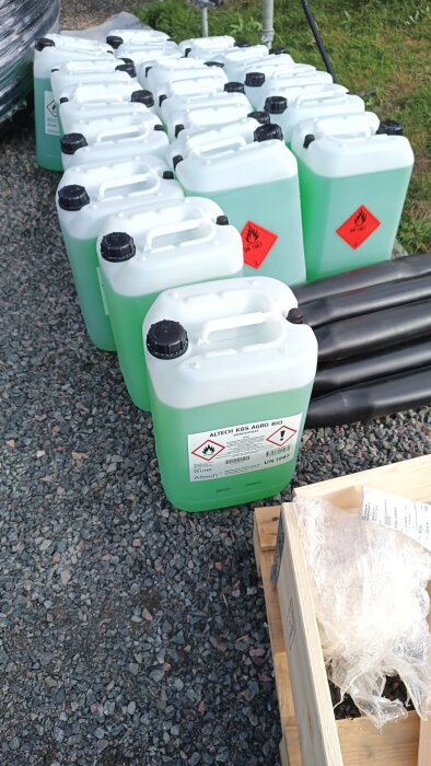 Rad av gröna plastdunkar med vätska, etiketterade med varningssymboler för brandfarliga material, står på grus.