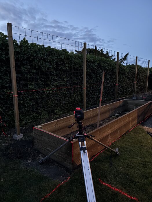 Odlingslåda under konstruktion med laserverktyg på stativ, omgiven av trädgård och insynsskydd i skymningen.