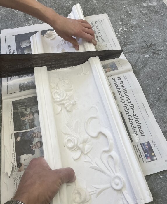 Dekorativ gipsskena med blommönster provas mot en bräda ovanpå tidningar. En hand justerar skenan.