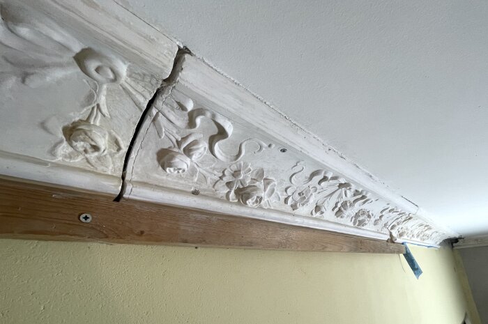Dekorativ taklist med blommönster gjord av gips, monterad över en träbräda. Synlig skarv i mitten av listen.