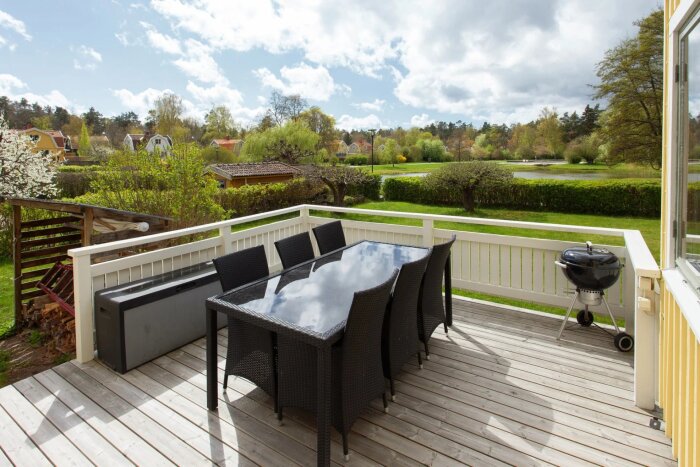 Uteplats med glasbord, åtta svarta stolar och grill, omgiven av staket och grönska, under en molnig himmel.