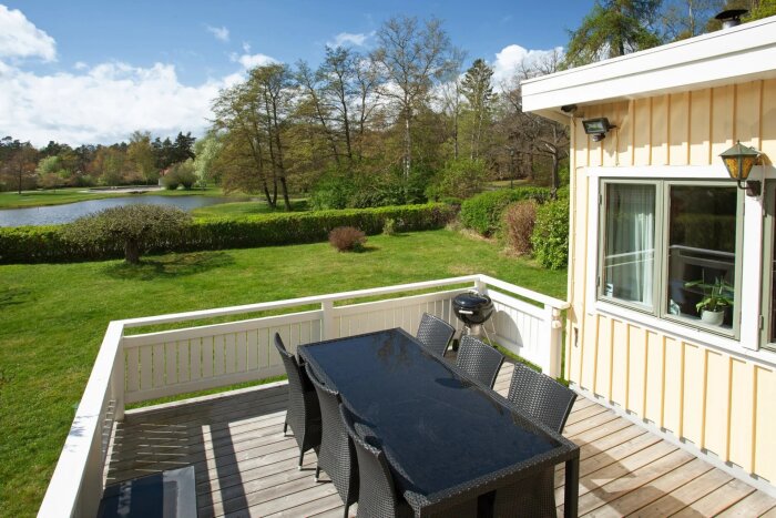 Uteplats med matbord och stolar i rotting, omgiven av trädgård med gräsmatta och träd i bakgrunden.