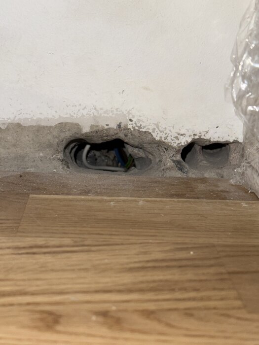 Öppning i väggen vid golvet med synliga kablar, delvis omgivna av flytspackel eller betongliknande material.