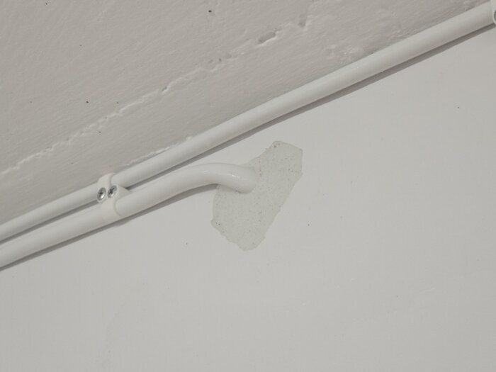 Närbild på en vit vägg där en bit puts har lossnat runt ett borrat eluttag, med en vit elkabel fäst i väggen.