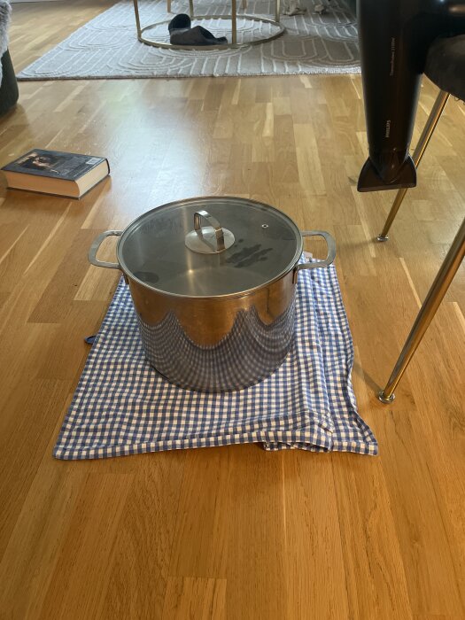 Stor kastrull med lock placerad på en blåvitrutig kökshandduk och disktrasa på trägolv.