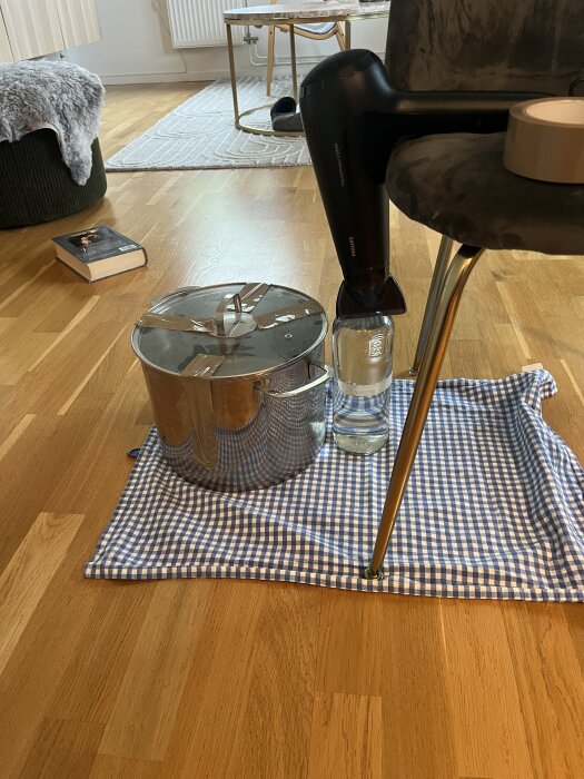 Hårfön blåser på en glasflaska och kastrull på golvet, stående på en blåvit-rutig duk med en stol som stöd i ett vardagsrum.