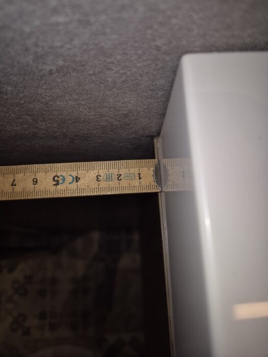 Måttstock ligger vinkelrätt över kanten av en vit kommod och mot en vägghörn i ett badrum, sedd från sidan med måttet 17 cm tydligt.
