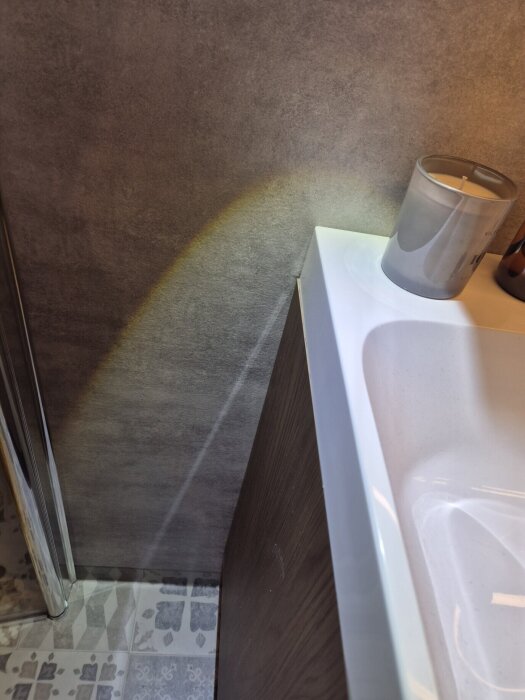 Badrumskommodens hörn och vägg syns, ett stearinljus står på kommoden bredvid handfatet. Kakel med mönster finns på golvet.