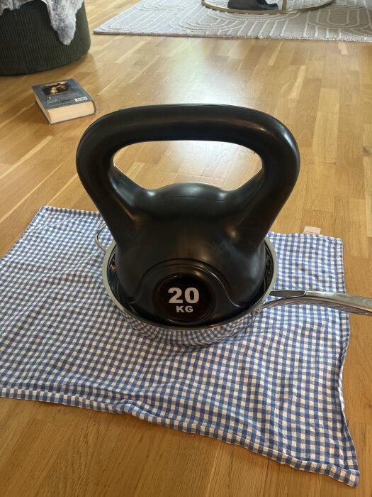 Kettlebell på 20 kg placerad i en kastrull på en blåvitrutig duk, med en bok i bakgrunden på ett trägolv.