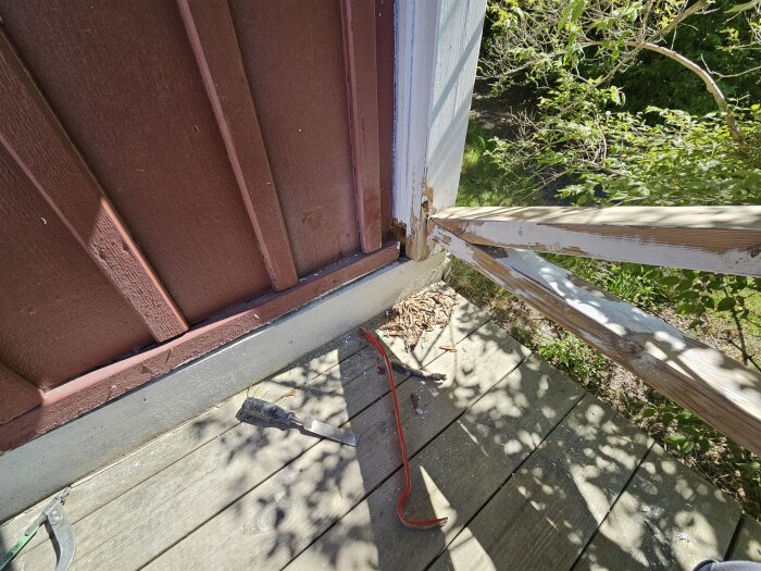 Skadat trä i husfasad med rötskador, en stämjärn ligger på trallen, omgiven av grönska; potentiellt arbete för att byta eller reparera trädetaljer.