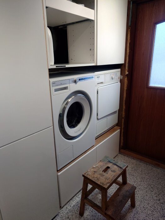 Tvättmaskin och torktumlare installerade i en vit förvaringsenhet bredvid en dörr, med en träpall framför.