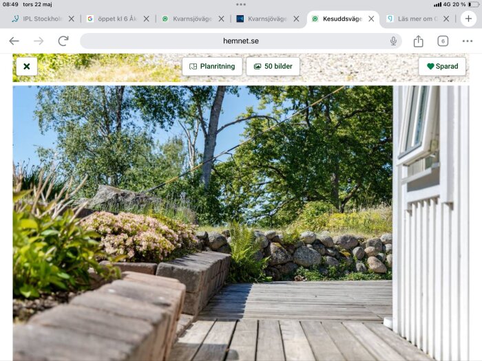 En lutande stenmur i en trädgård med blommor, en träterrass och en vit husvägg syns intill skogsklädda träd under en klar himmel.