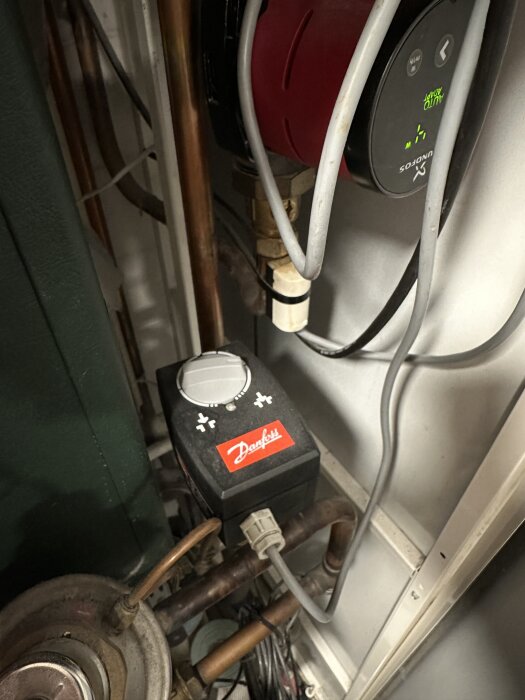 Danfoss ECL 100M regulator med vridknapp, placerad bland rör och kablar i ett fjärrvärmesystem.