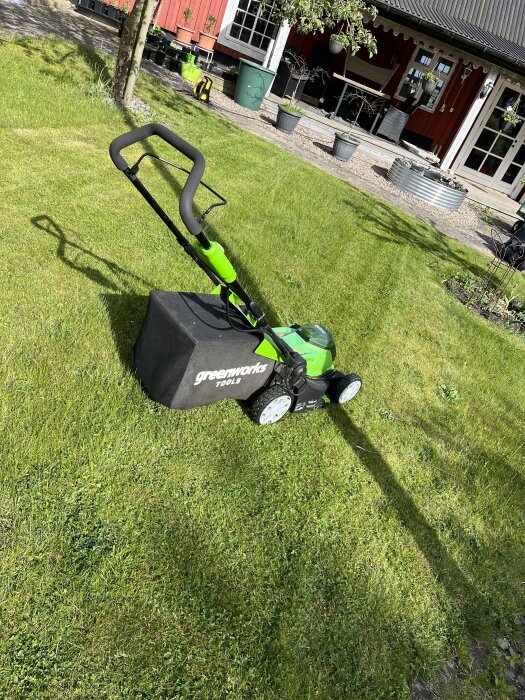 Gräsklippare med grönt handtag står på en halvklippt gräsmatta framför ett rött hus med vita fönster och flera krukväxter.