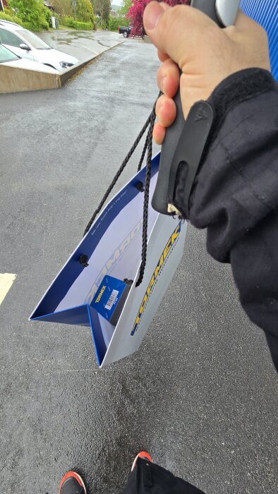 En person håller en papperspåse från Tormek utomhus. Påsen innehåller en TT-50 slipjig. Asfalt och parkeringsplats syns i bakgrunden.