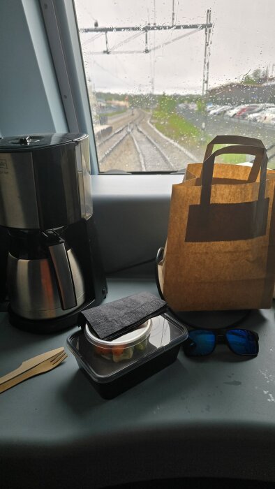 Matsäck med sallad, kaffetermos och solglasögon på ett tåg med regnig utsikt genom fönstret.