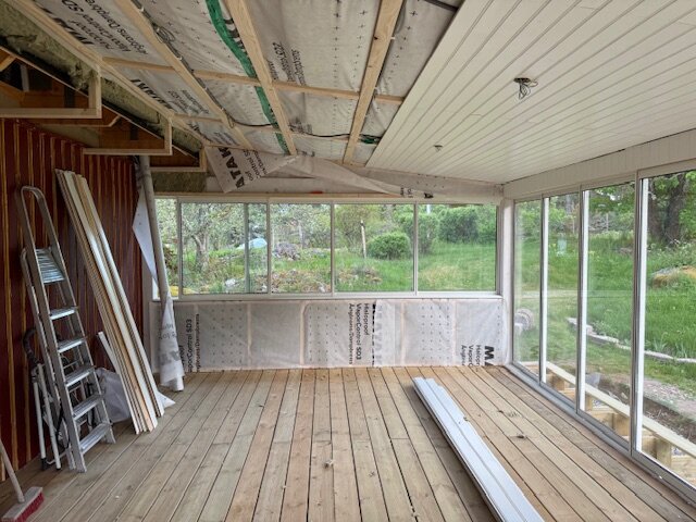 Isolerat uterum under byggnation med trätak och fönster längs ytterväggarna, samt material upplagt på golvet. Stege och byggmaterial står längs en vägg.