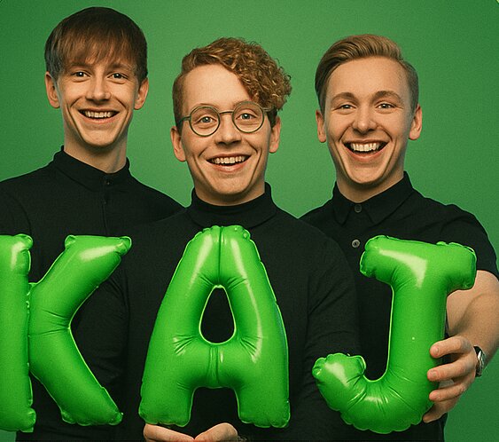 Tre glada personer håller upp gröna ballongbokstäver som bildar ordet "KAJ" framför en grön bakgrund.