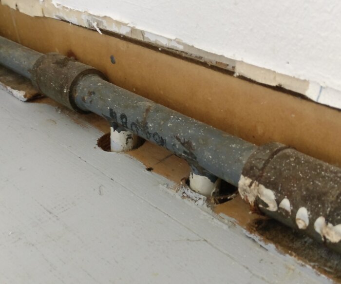 Gammalt rör med skarvkoppling vid golv och vägg; sliten yta och påverkan av tid.