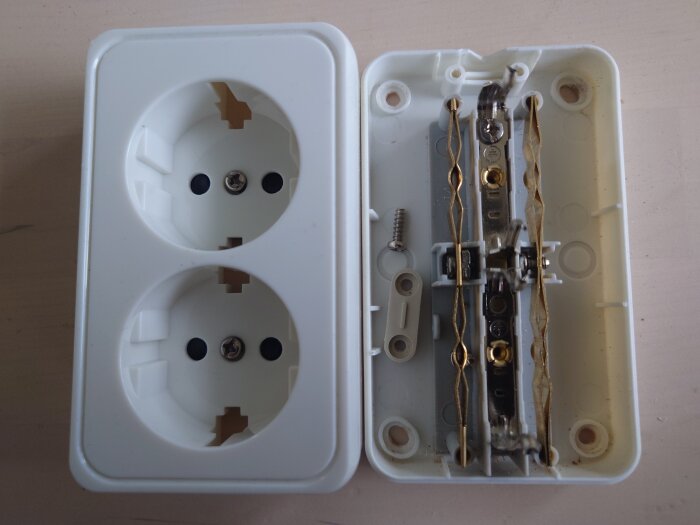Öppet vägguttag från Malmbergs, vit plast med två uttag och synlig metalldel på insidan, ligger på träyta.