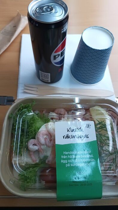 Räksmörgås i plastförpackning med etikett, burk Pepsi Max, pappersmugg och träbestick på bord.