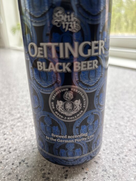 Närbild på en OeTTINGER Black Beer-burk med blå dekorationer, placerad på ett grått köksbänkskiva.